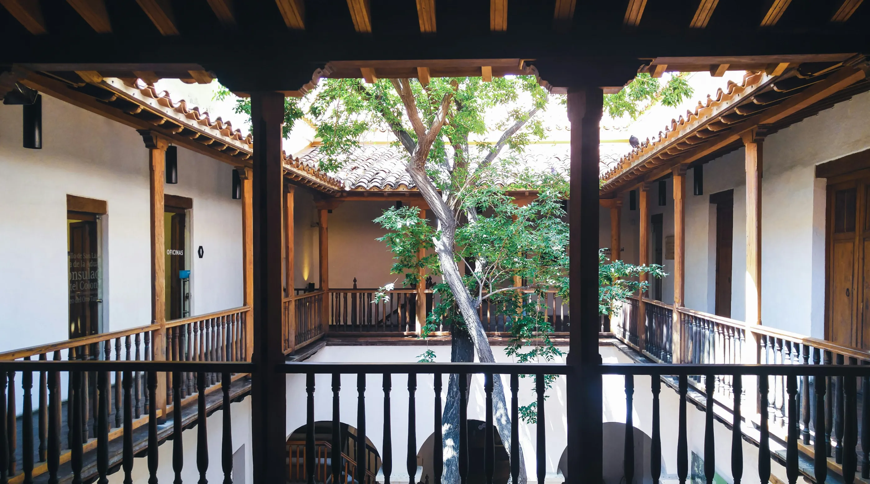 Patio colonial con balcones de madera y un árbol al centro, con luz que entra desde arriba